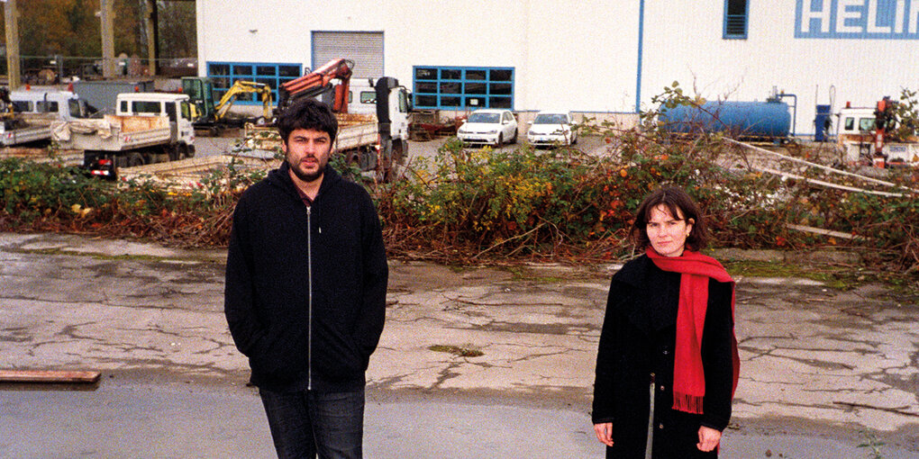 Edis Ludwig und Emily Wittbrodt, die das Duo Ludwig Wittbrodt, stehen vor einem Transportgelände. Hinter ihnen sind eine Halle mit weißen Außenwänden aus Metall sowie diverse Klein-LKWs zu sehen.