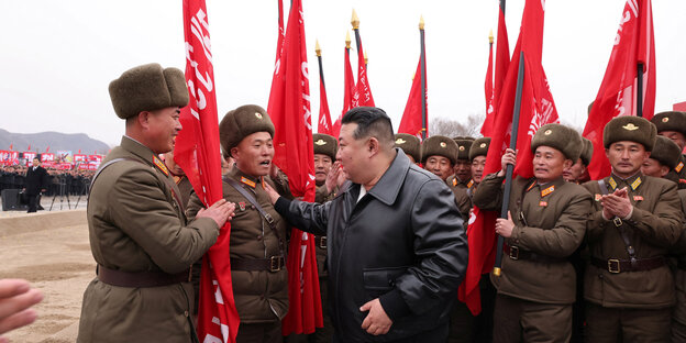 Nordkoreas Machthaber Kim Jong Un zwischen Männern in braunen Wattejacken und mit roten Fahnen