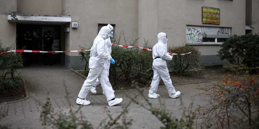 Drei Personen in weißen Schutzanzügen vor einem Mietshaus.