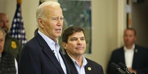 Joe Biden, ein alter Mann mit weißen Haaren redet. Er trägt einen dunklen Anzug und ein helles Hemd, ebenso tun das zwei drei Personen im Hintergrund. Auch im Hintergrund: eine Fahne der USA, ein Mensch in Uniform und ien Mensch in dunklem Pollunder.