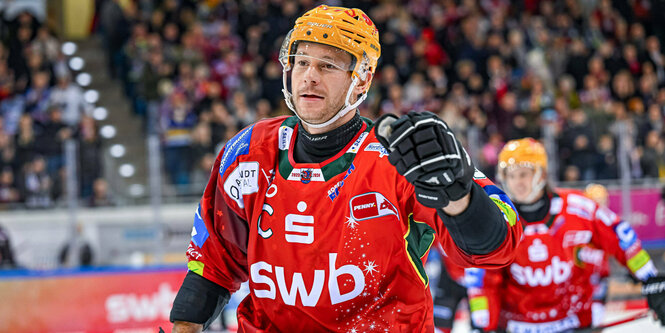 Jan Urbas während einem Spiel. Er trägt ein rotes Trikot und einen Orangenen Helm. Seine linke Hand, in Handschuh, hat er erhoben. Im Hintergrund Publikum und ien weiterer Spieler auf demEis.