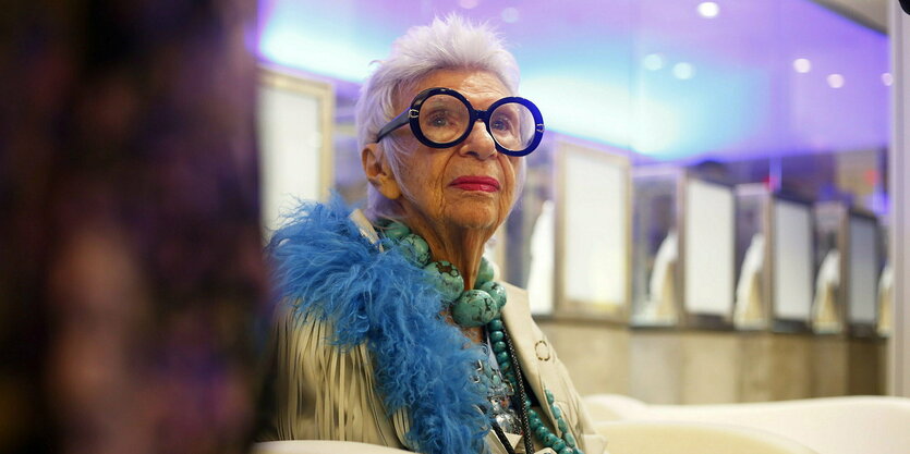 Iris Apfel sitzend, schmuckbehangen, mit blauer Fedeboa und schwarzer Brille