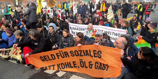 Viele Demonstrierende sitzen auf einer Straße und blockieren diese. Sie halten verschiedene, bunte Fahnen in die Luft, unter anderem Regenbogenflaggen. In den Händen halten sie ein Transparent, auf dem sie fordern: "Geld für Soziales, statt Kohle, Öl und Gas!"