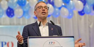 Mann hält am Pult Rede, hinter ihm blaue Luftballons