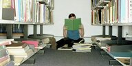 Eine Person sitzt in einer Bibliothek auf dem Boden zwischen Bücherregalen und liest