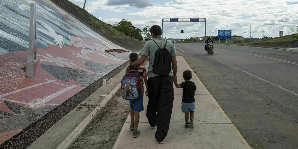 Ein Mann und zwei Kinder von hinten auf einem Bürgersteig neben einer Straße. Der Mnn hat einen Arm um das eine Kind gelegt und hält das andere an der Hand.