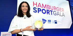 Reem Khamis bei der Ehrung zu Hamburgs Sportlerin des Jahres. Sie trägt offene, schulterlange schwarze Haare, ein weißes Shirt mit modisch-weiten Ärmeln und in der linken Hand den Preispokal: Eine Art runde Scheibe, durch die ein stilisiertes Hamburger Rathaus hindurchscheint.