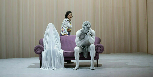 Auf einem violetten Sofa sitzt eine Darstellerin unter einem Schleier, daneben eine sitzende Herkules-Skulptur, dahiner eine weitere Darstellerin