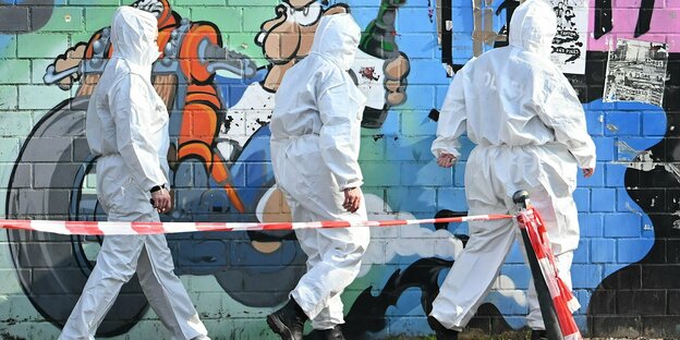 Drei Personen in weißen Schutzanzügen vor einer mit Graffiti besprühten Mauer