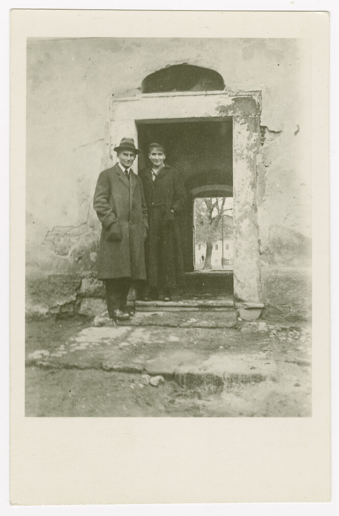 Schwarz-Weiß-Foto von Franz Kafka (rechts im Bild) und Ottla Kafka (links). Die beiden Geschwister stehen vor der Tür eines alten Hofs, die Wände sind teils abgebröckelt. Franz Kafka trägt einen Mantel, Krawatte und Hut. Ottla Kafka trägt einen dunklen Mantel mit Nadelstreifen.