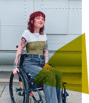 Luisa L'Audace ist im Rollstuhl vor einer mintgrünen Wand zu sehen. Sie trägt eine Jeans und ein Shirt, auf ihren Armen sind viele Tattoos, unter anderem von Frida Kahlo mit Zigarette