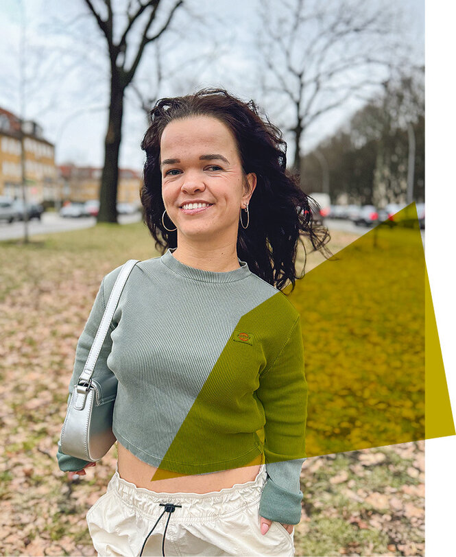 Portrait von Janina Nagel, die mit grünem Oberteil und Handtasche vor einer Wiese steht, im Hintergrund sieht man einen kahlen Baum und eine Häuserzeile