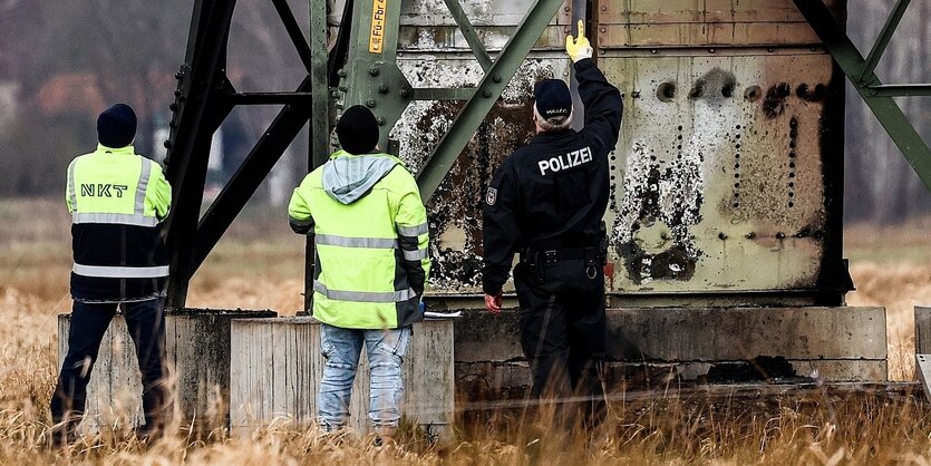 Drei Männer stehen vor einem Strommasten mit Brandspuren