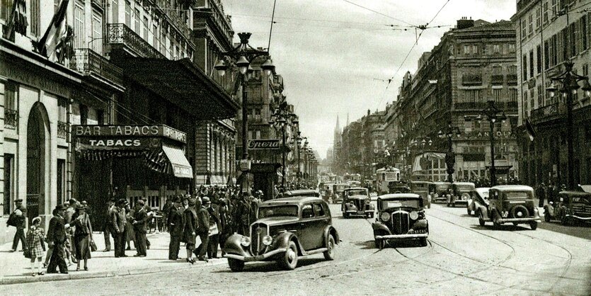 Straßenszene aus Marseille 1940, Autos fahren auf der Straße mit Straßenbahngleisen, im Hintergrund ist ein "Tabac" zu sehen, Menschen auf dem Gehweg