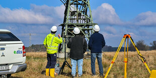 Drei Arbeiter vor einem Strommast. Im Hintergrund ein Feld. Links neben den Arbeitern ein Auto. Rechts neben ihnen ein Gerät zur Vermessung.