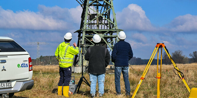 Drei Arbeiter vor einem Strommast. Im Hintergrund ein Feld. Links neben den Arbeitern ein Auto. Rechts neben ihnen ein Gerät zur Vermessung.