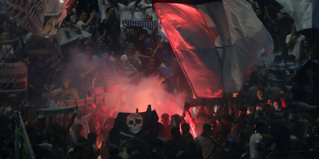 Fankurve in einem Stadion. Die Fans schwenken Fahnen. Ein rotes, bengalisches Feuer leuchtet.