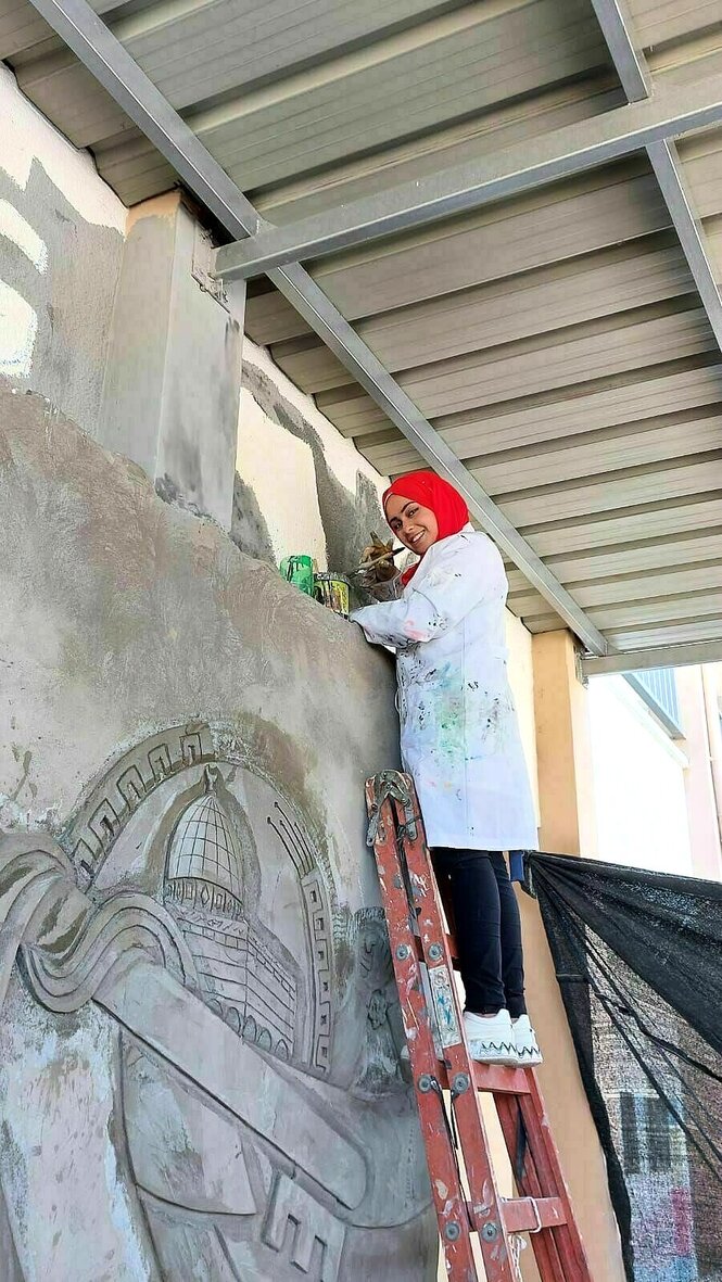 Ein Bild aus glücklchen Tagen: Menna Hamouda steht auf einer Leiter mit Malerkittel und rotem Kopftuch und lächelt in die Kamera