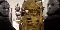 Eine goldene Maske, der Raubkunst aus Benin nachgebildet, im Zentrum. Links und rechts zwei Personen mit weißen Masken