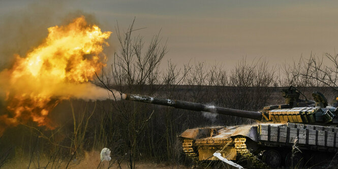 Rechts ein Panzer, der nach Links feuert. Man sieht das Feuer sogar. Im Hintergrund Sträucher und karge Landschaft. Alles ist gräulich.