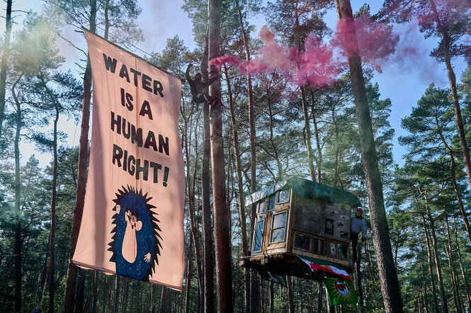 Blick in den besetzten Wald in Grünheide, vor dem Baumhaus hängt ein Banner: Water is a human right! Auf einem Baum schwenkt jemand eine brennende rote Fackel