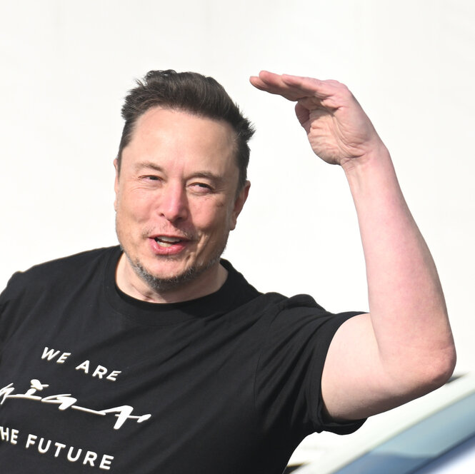 Ein gut gelaunter Elon Musk winkt, bevor er das Betriebsgelände verlasst. auf seinem T-Shirt steht: We are Tesla - the future