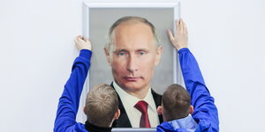 Zwei Arbeiter in blauer Arbeitskleidung hängen ein Porträt des russischen Präsidenten Wladimir Putin auf