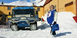 Ein weibliches Mitglied der Wahlkomission steht mit einer tragbaren Wahlurne neben einem blauen Schneeräumer auf einem schneebedeckten Platz