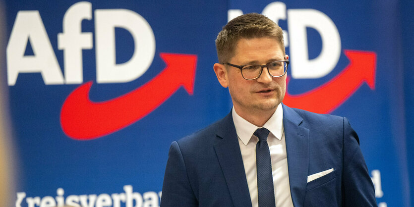 Blonder Mann mit Brille, Anzug und Krawatte, vor blauem Hintergrund mit AfD-Motiven