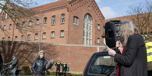 Eine Frau spricht in ein Mikrofon, im Hintergrund eine rote Gefängnismauer