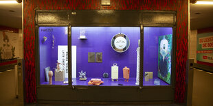 Blick in eine Fenstervitrine auf einem U-Bahnsteig. Die Wände der Vitrine sind in strakem Lila ausgekleidet. In der Vitrine sind zahlreiche Skulpturen und Wandarbeiten zu sehen.