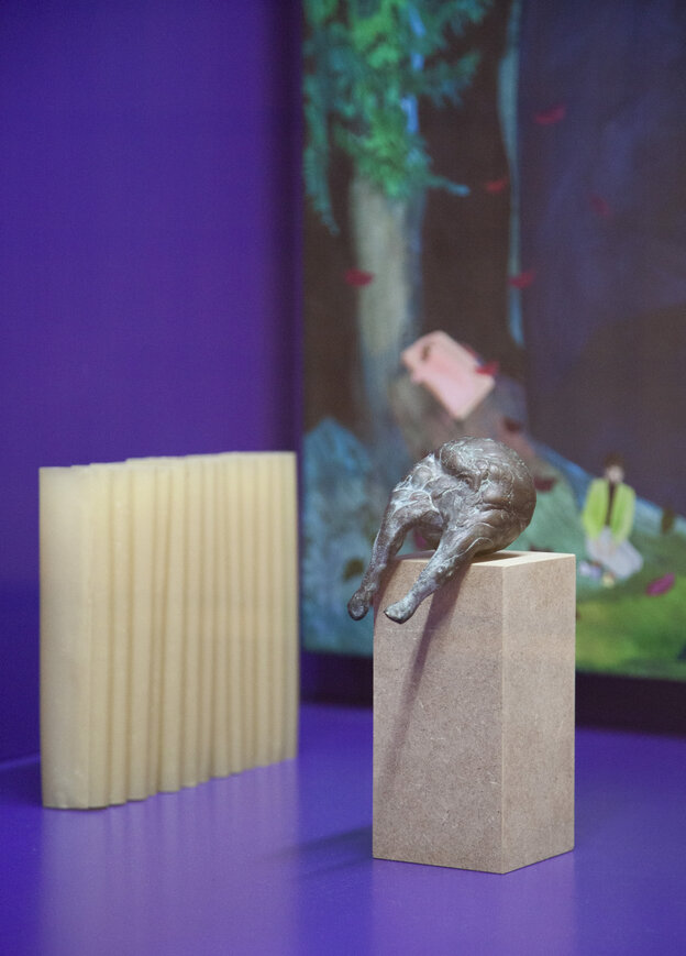 Eine Skulptur aus Metall hat eine runde organische Form. Am unteren Teil der Skulptur scheinen sich Beine zu formen. Sie ist auf einen Holzklotz aufgesetzt. Dahinter sind eine beige Vase mit Ziehharmonika-Falten und die untere Ecke eines Gemäldes mit eine