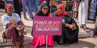 3 Frauen mit Kopftüchern sitzen auf dem boden und halten ein Schild, um gegen die Genitalverstümmelung zu demonstrieren