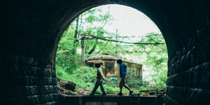 Blick durch ein Tunnel: Zwei Jungen gehen hintereinander, im Hintergrund ein verlassener Eisenbahnwagen