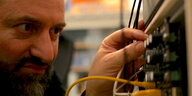 Der Musiker Jörg Schweinbenz dreht an den Knöpfen eines Musikapparats, aus dem bunte Kabel abstehen. Schweinbenz trägt einen Bart.