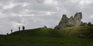 Burg auf einem grünen Hüge, menschen stehen herum und schauen in die Landschaft