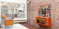 Ein Raum mit brauner Wand, ein oranges Regal an der Wand, darüber ein Gemälde mit einer Frau im Anzug, links im Bild ein Bürotisch, im Hintergrund ein Schaufenster, das auf die Straße blickt.