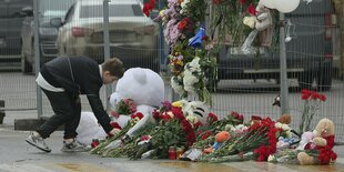 Ein Junge legt Blumen nieder vor einem Zaun, an dem schon viele Blumen und ein riesiger weißer Stoffbär liegen