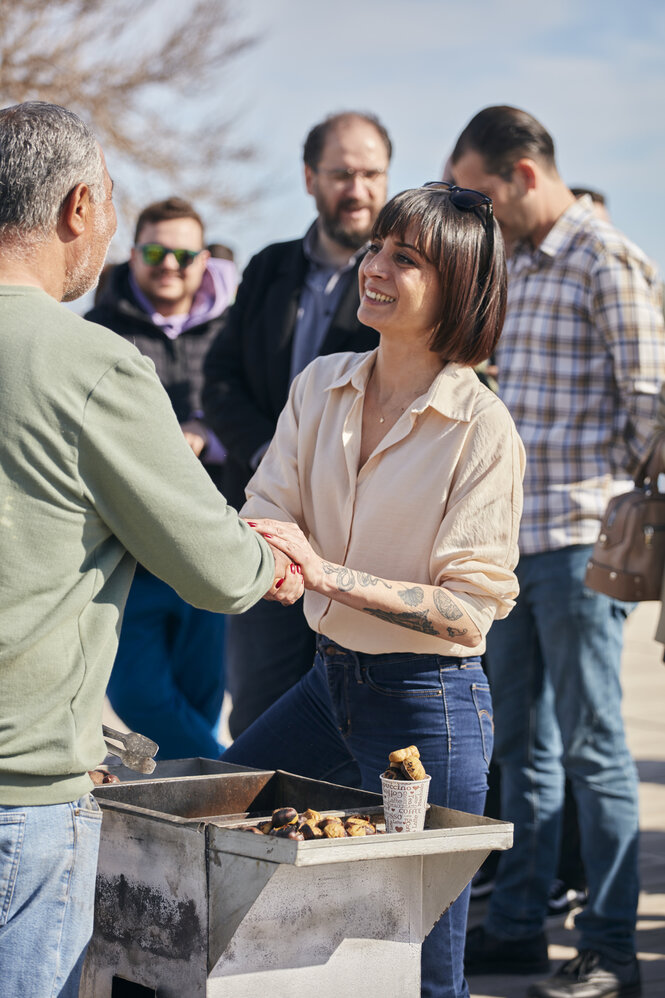 Eien junge Frau mit tätowierten Armen und hochgeschlagenem Hem fasst einen Mann an, der Kastanien grillt