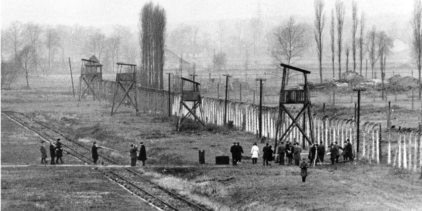 Menschen bewegen sich in einer düsteren Landschaft auf einen KZ-Eingang zu