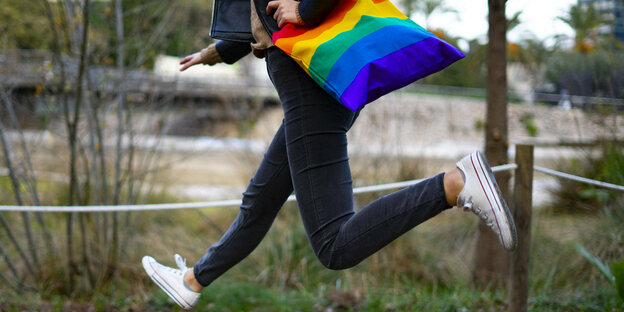 Junge Person hüpft mit einer Tasche in Regenbogenfarben auf einer Wiese