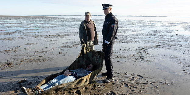 Zwei Männer tragen eine Leiche am Strand.