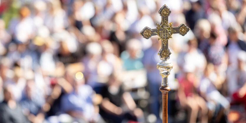 Ein Kreuz im Vordergrund, im Hintergrund Menschen unscharf