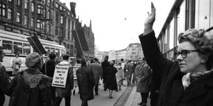 Schwarz-weiß-Foto, eine Frau steht mit erhobener Hand am Rande einer Demonstration. Die Teilnehmer wurden von hinten fotografiert - einer trägt ein Schild auf dem Rücken: Keine Bombe ohne Test - kein Test ohne Tod