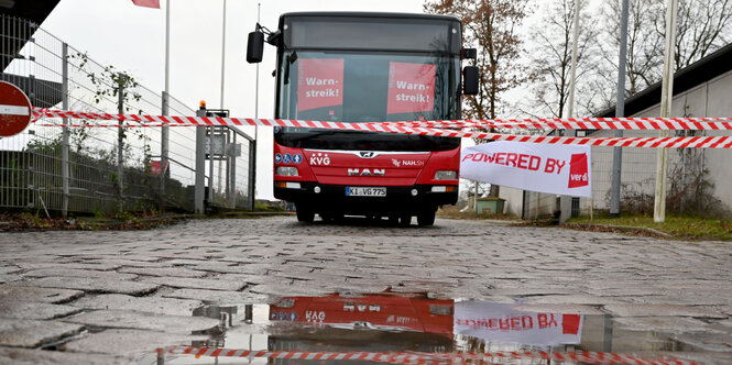 Ein roter Bus steht hinter einer roten Absperrung. Am Bus hängen Plakate, die zum Streik aufrufen. Im Vordergrund eine Pfütze.