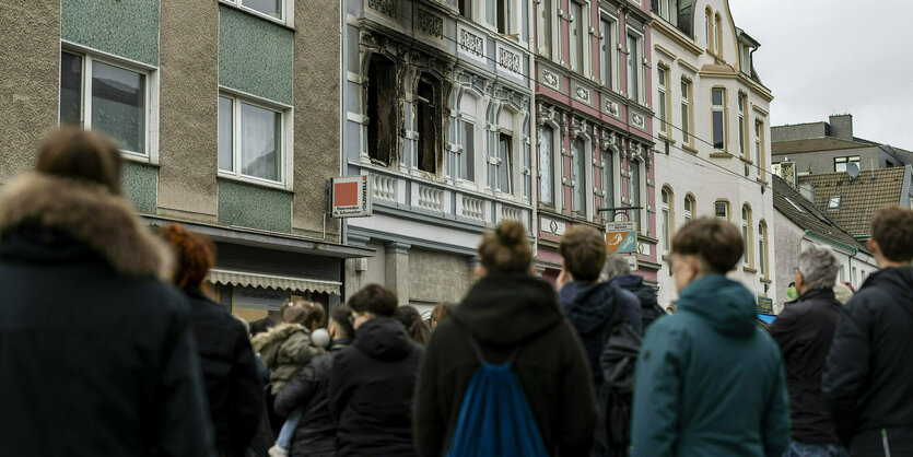 Menschen vor einem Haus, an dessen Fenstern Brandspuren zu erkennen sind