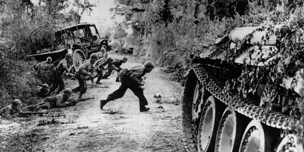 Soldaten rennen sich duckend über eine unbefestigte Straße, vorne steht ein Panzer, hinteer ihnen liegt ein Lastwagen im Graben