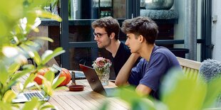 Zwei junge Männer sitzen auf einer Terasse hinter Laptops.