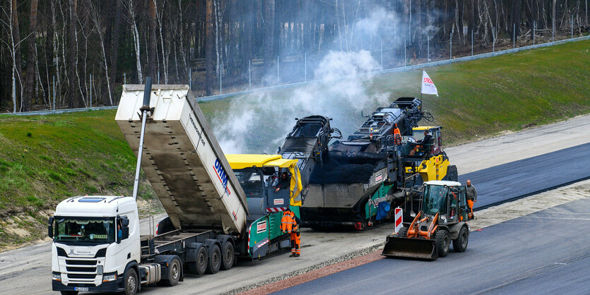 Arbeiter in Warnkleidung asphaltieren neue Autobahnstreifen am Rand eines Waldes mit einem Laster, einem Asphaltmischer und einem kleinen Bagger