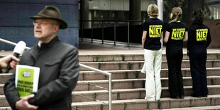 Drei protestierende junge Frauen auf einer Treppe, die mit dem Rücken zur Kamera stehen und auf deren T-shirt steht: We kunnen niet wachten (wir können nicht warten) Im Vordergrund wird ein Mann interviewt